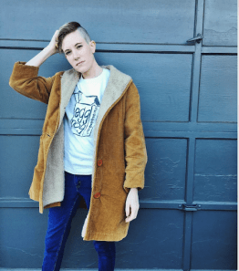 Michelle stands in front of what looks like a factory door, wearing a large coat with jeans and a t-shirt that says, “eggnog skateboards”. They’re running their hands through their hair with their right arm.